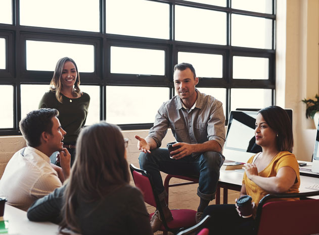Grupo de cinco personas conversando en una sala con ventanales, una de ellas de pie